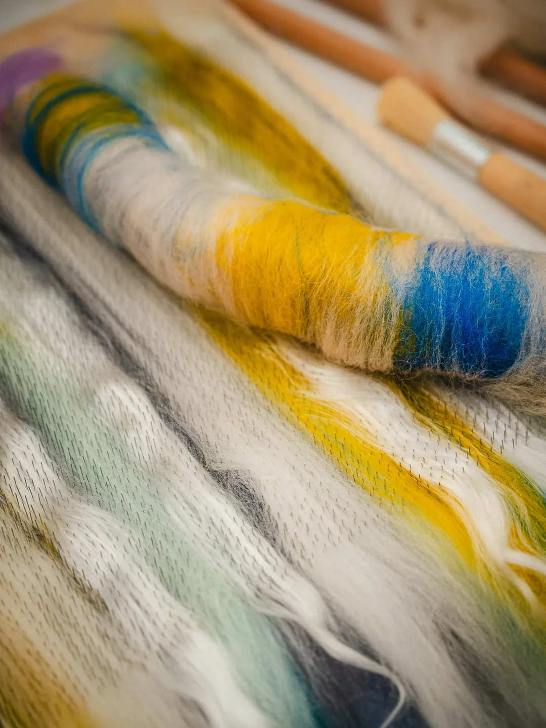 Mecha de lana teñida en azul y amarillo sobre una lámina de prefieltro, con herramientas de madera al lado